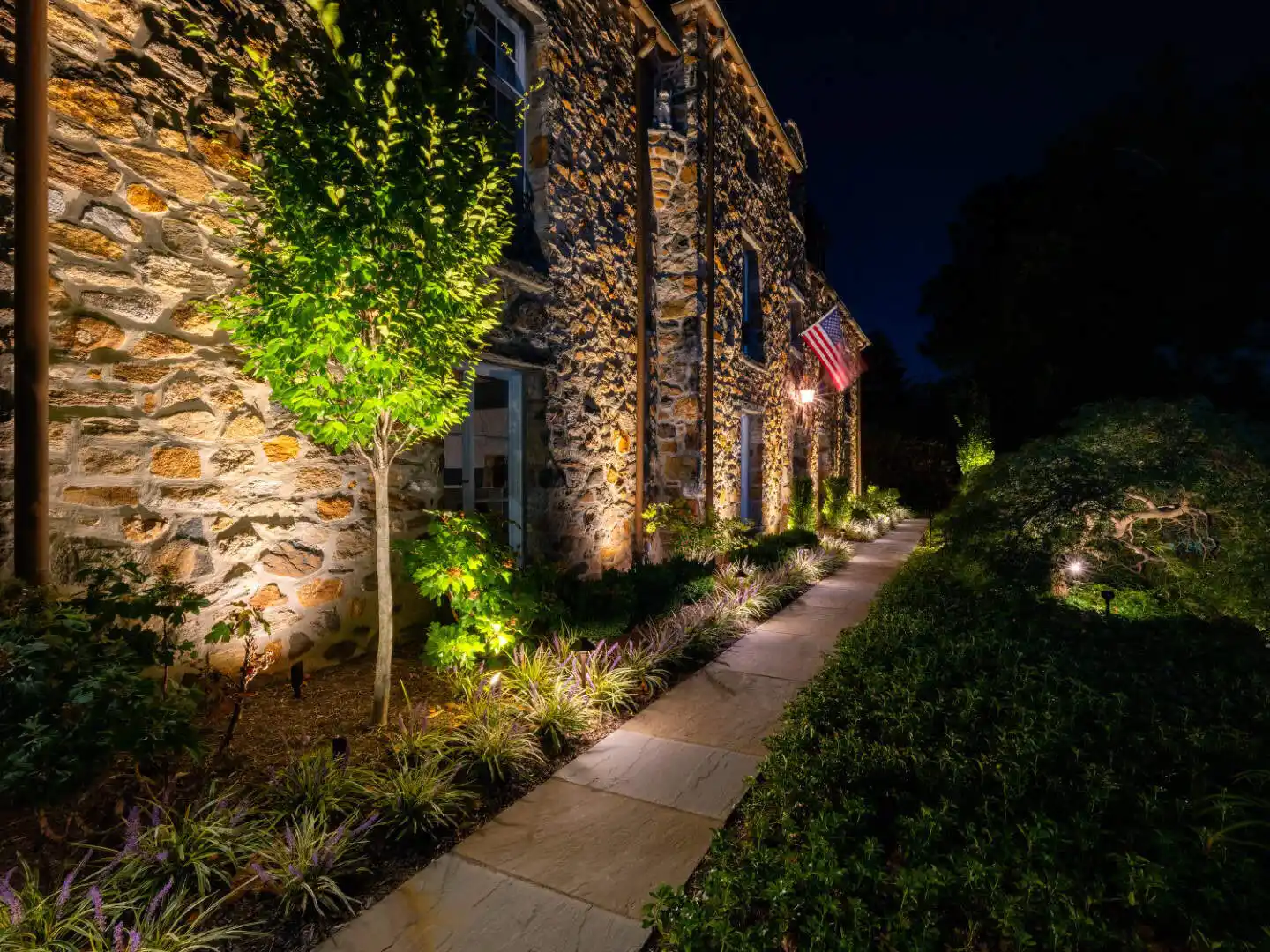Stone house exterior at night, with warm lighting accentuating textured walls. A well-lit pathway with plants and artful hardscaping leads alongside the building. An American flag is displayed near the entrance, while expertly manicured bushes line the way—showcasing top-notch landscaping services in Delaware County PA.
