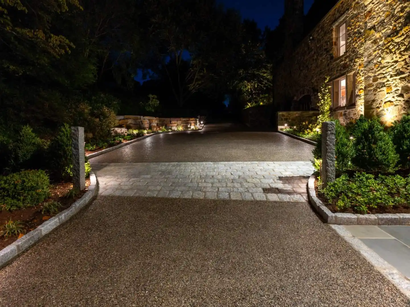 The illuminated driveway at night leads to a stone house, its warmly lit windows inviting you in. Expert landscaping services in Delaware County, PA have lined the path with green shrubs and accented it with stone columns against a backdrop of silhouetted trees.