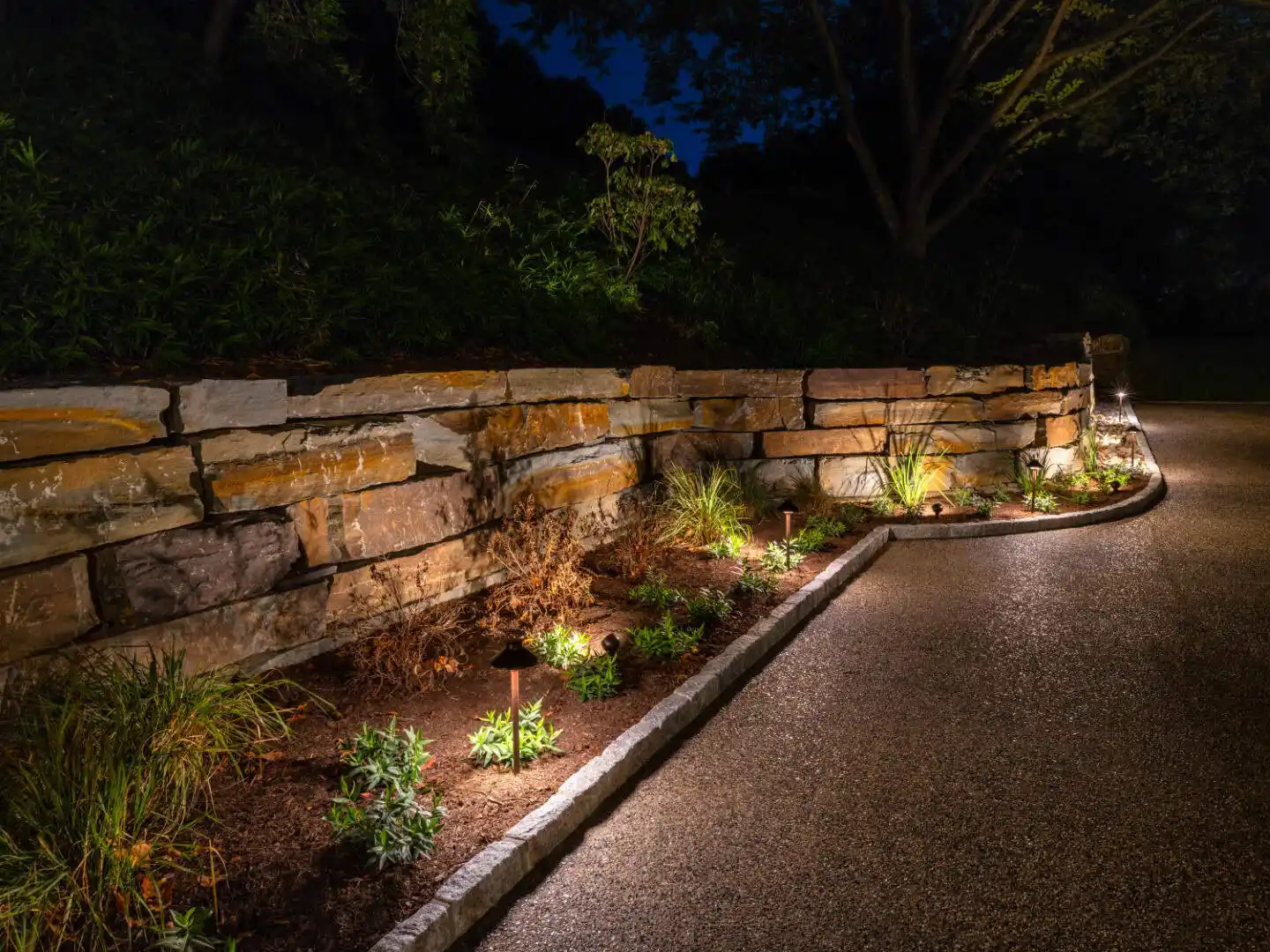 A curved stone wall with a garden bed is beautifully lit by small landscape lights at night. The paved path beside it invites exploration, while trees stand silhouetted against the dark sky, showcasing expert hardscaping services in Delaware County, PA.