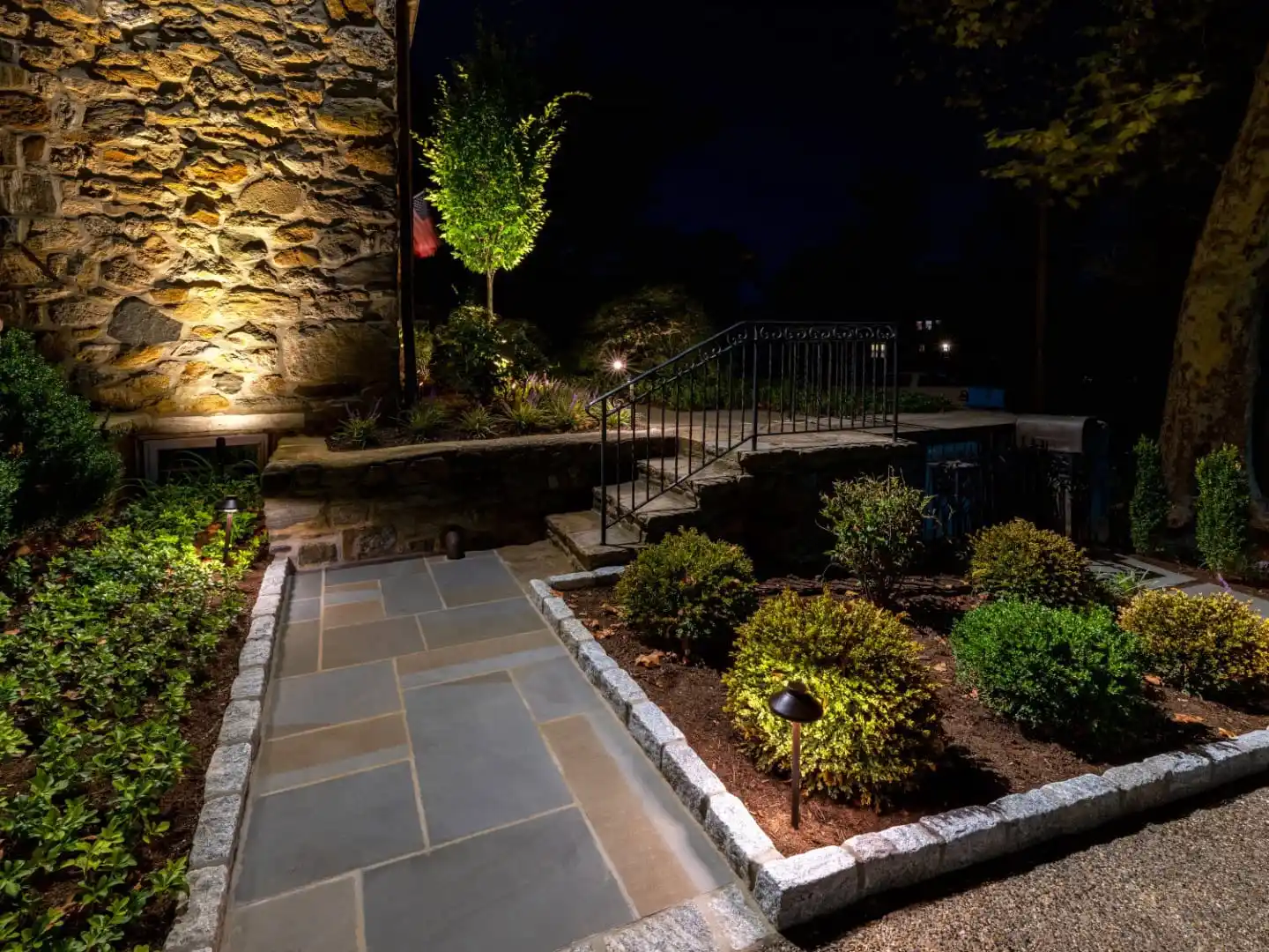 A nighttime garden scene with a stone walkway surrounded by neatly trimmed bushes and shrubs showcases the beauty of outdoor living in Delaware County, PA. A small staircase with a metal railing leads to a raised area. Ambient lighting highlights the stone wall and a small tree.