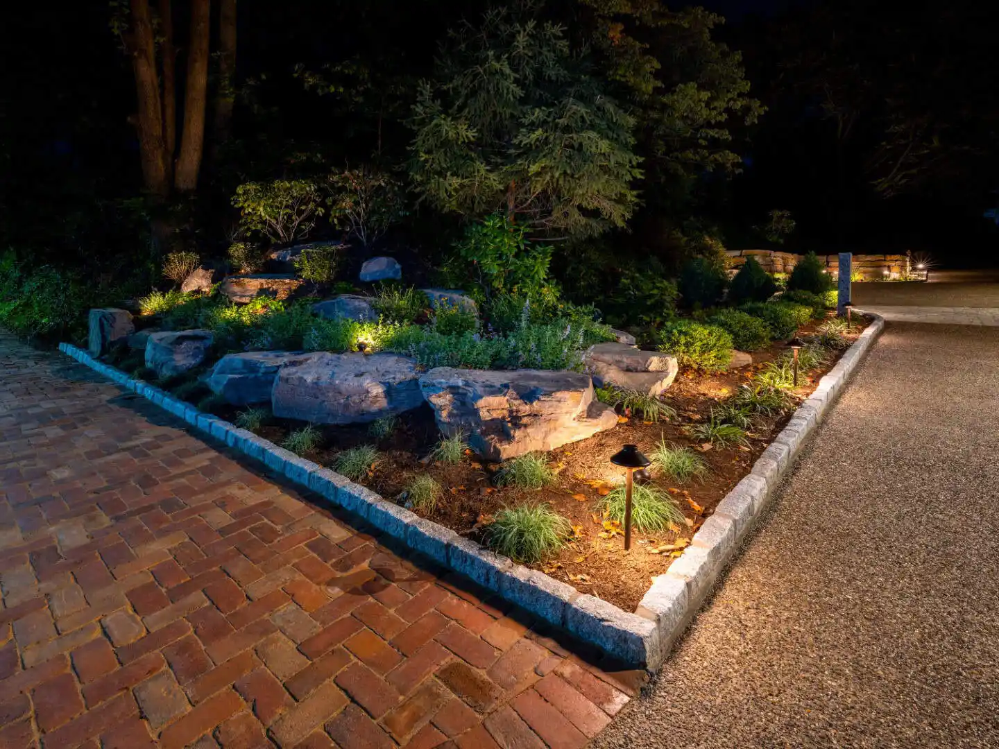 A landscaped garden at night with spotlights illuminating rocks and plants is bordered by a stone edge, adjacent to a brick pathway. Trees and shrubs provide a dark contrast in the background, showcasing the beauty of outdoor living in Delaware County, PA.