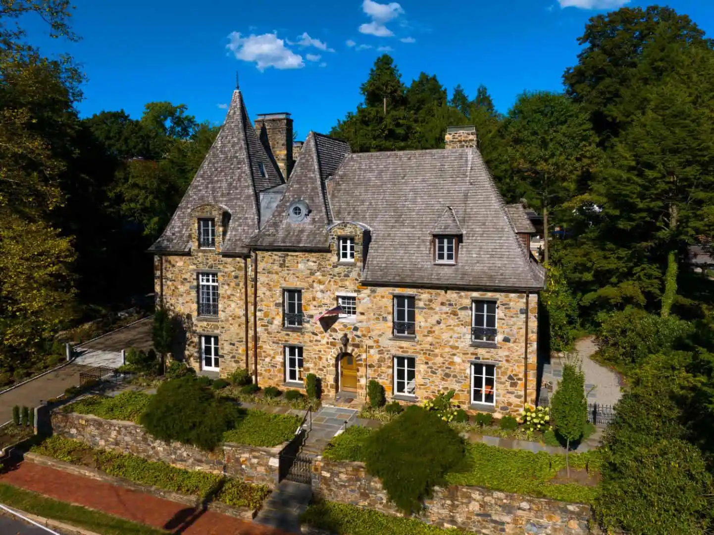 A historic, multi-story stone house with a steep, pointed roof and dormer windows stands proudly. An American flag adorns the entrance. Surrounded by lush trees and expert hardscaping services in Delaware County, PA, the building sits behind a low stone wall under a sunny sky.