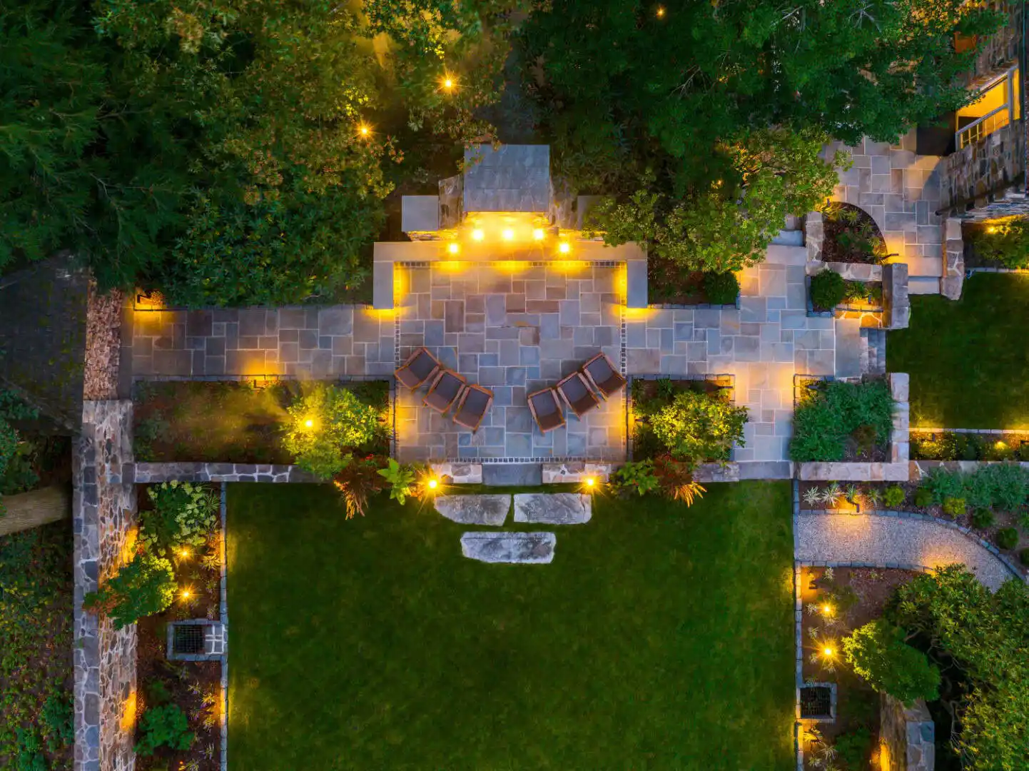 Aerial view of a beautifully lit patio and garden at night. The stone patio features wooden chairs and glowing lights outlining the area. Expert hardscaping services in Delaware County, PA have enhanced the lush green grass and pathways, with trees and plants providing a natural border.