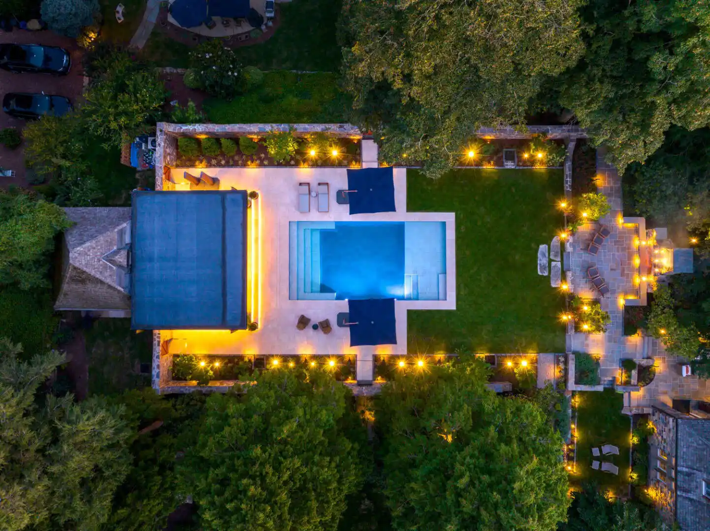 The illuminated driveway at night leads to a stone house, its warmly lit windows inviting you in. Expert landscaping services in Delaware County, PA have lined the path with green shrubs and accented it with stone columns against a backdrop of silhouetted trees.