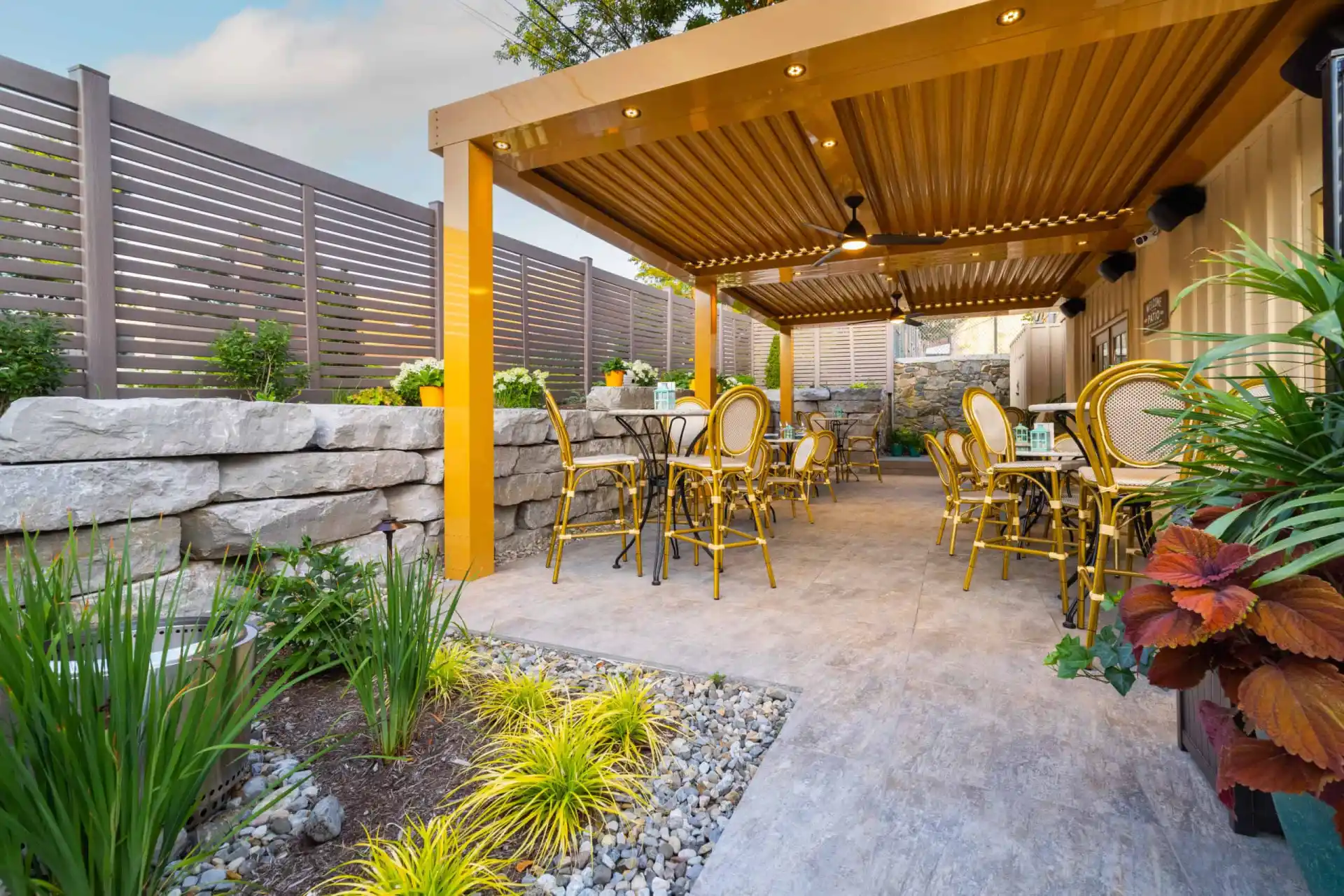 A cozy outdoor patio with yellow chairs and tables nestles under a wooden pergola, offering delightful outdoor living in Delaware County, PA. Bordered by stone walls and lush greenery, slatted fencing ensures privacy while colorful flowers bloom beneath a partly cloudy sky.