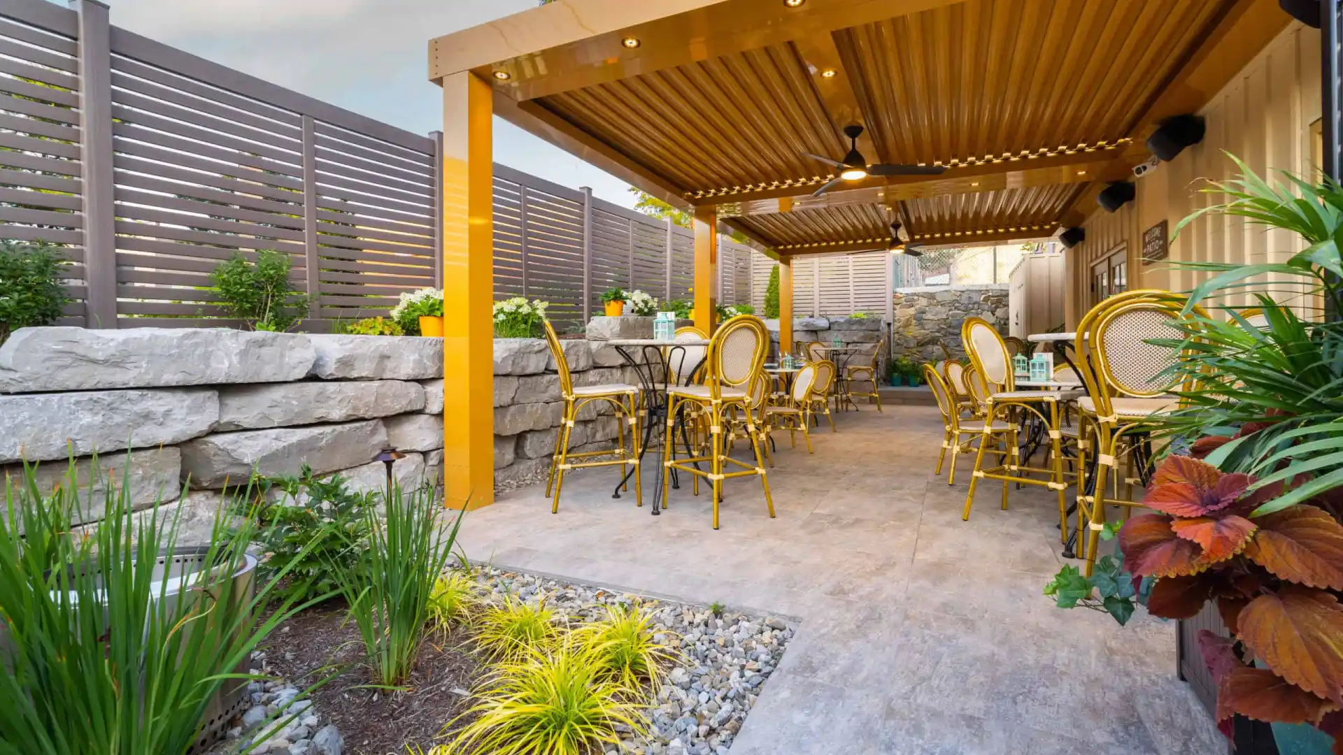 A cozy outdoor patio with yellow chairs and tables nestles under a wooden pergola, offering delightful outdoor living in Delaware County, PA. Bordered by stone walls and lush greenery, slatted fencing ensures privacy while colorful flowers bloom beneath a partly cloudy sky.