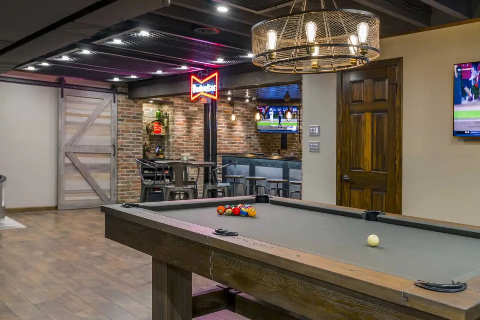 A cozy game room with a pool table takes center stage. In the background, a seating area offers a table and chairs, accented by a neon sign and wall-mounted TV. Rustic wooden and brick decor evoke outdoor living charm, while a circular chandelier casts a warm glow from above.
