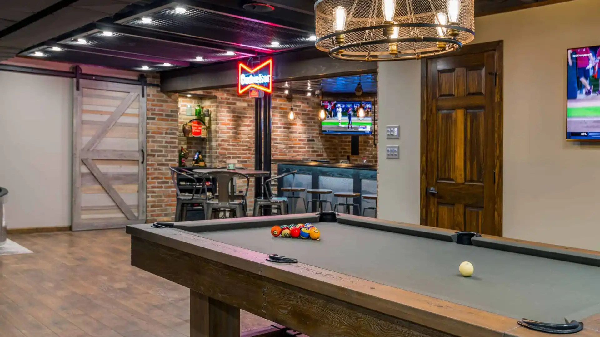A rustic room in Delaware County, PA, features a billiards table, a glowing neon sign, and a TV above a cozy bar area with stools. Wooden accents and a chandelier infuse warmth into the modern design, reminiscent of the region's refined outdoor living ambiance.
