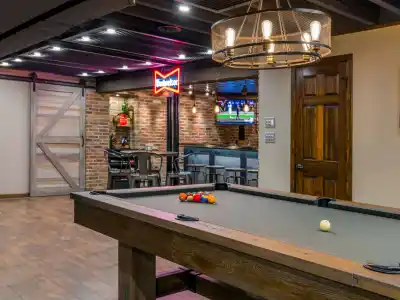 A rustic room in Delaware County, PA, features a billiards table, a glowing neon sign, and a TV above a cozy bar area with stools. Wooden accents and a chandelier infuse warmth into the modern design, reminiscent of the region's refined outdoor living ambiance.