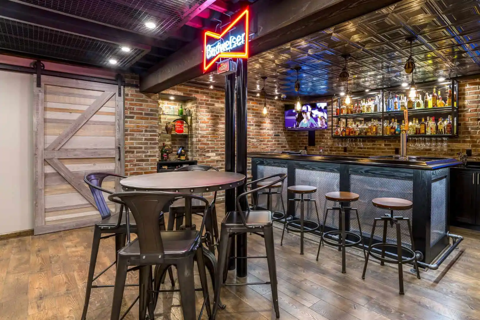 A cozy bar with a wooden floor and brick walls features high tables with stools, a stocked bar boasting colorful bottles, and a neon 