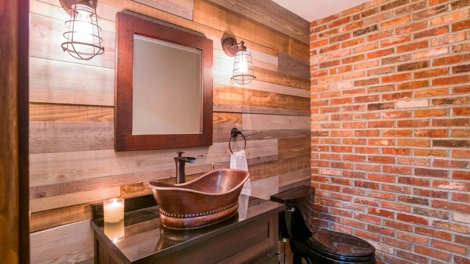 Rustic bathroom with a wooden vanity and copper vessel sink. A rectangular mirror hangs above, flanked by industrial-style fixtures. The walls feature wood paneling, exposed brick, and subtle touches reminiscent of bespoke outdoor living in Delaware County, PA. A lit candle sits on the counter.