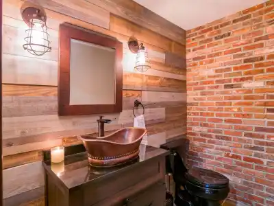 Rustic bathroom with a wooden vanity and copper vessel sink. A rectangular mirror hangs above, flanked by industrial-style fixtures. The walls feature wood paneling, exposed brick, and subtle touches reminiscent of bespoke outdoor living in Delaware County, PA. A lit candle sits on the counter.