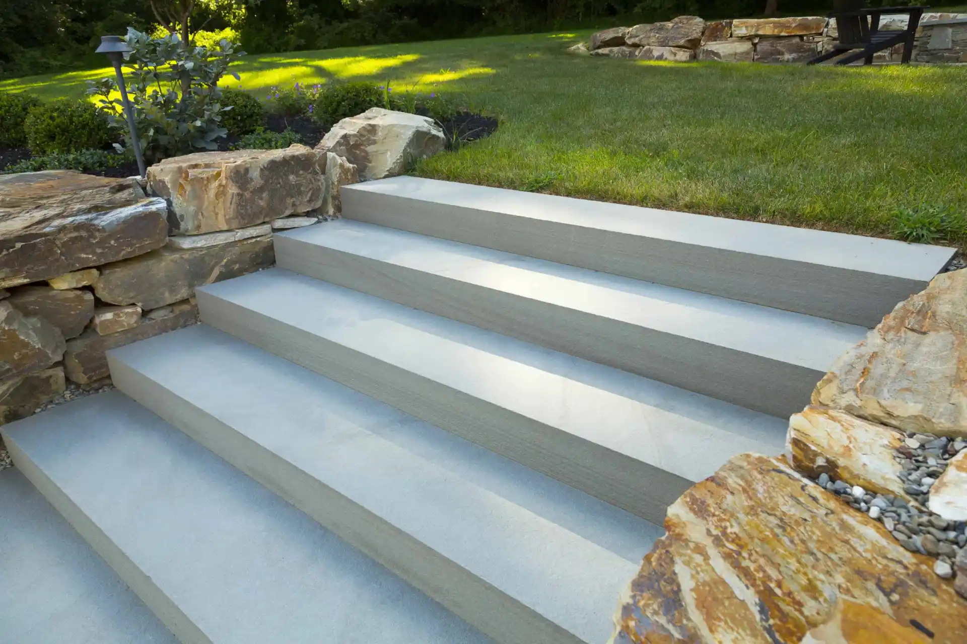 Wide concrete steps bordered by large natural stone walls lead up to a grassy lawn, with landscaping and greenery visible in the background.