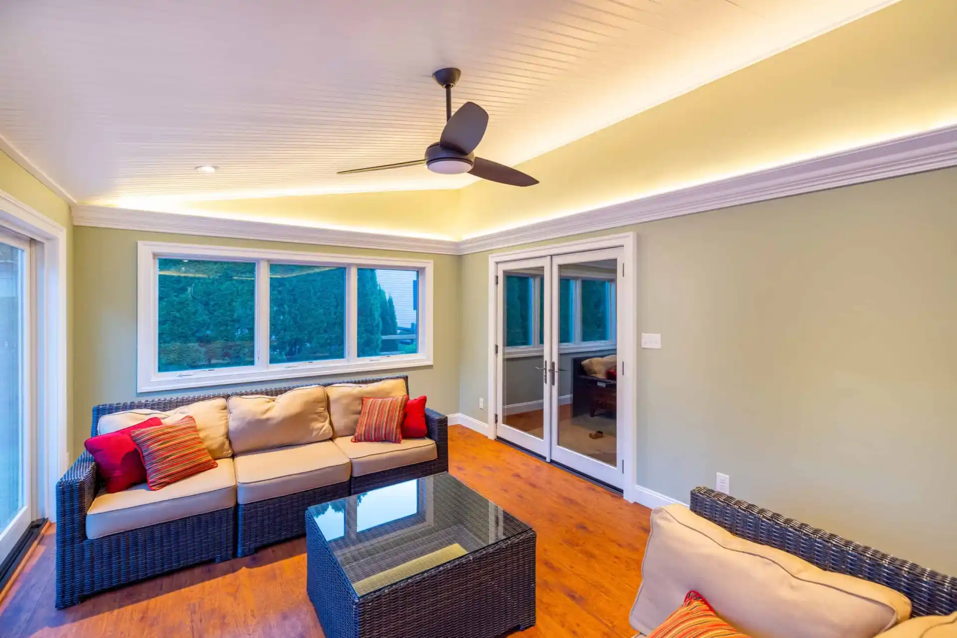 A cozy sunroom with wicker furniture featuring beige cushions and red accent pillows invites relaxation, while a glass-top coffee table anchors the space. Large windows and sliding glass doors offer abundant natural light, seamlessly connecting to the outdoor living area in Delaware County PA.