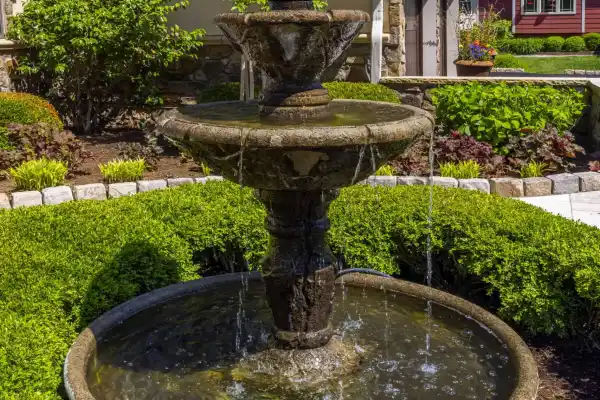 A tiered stone fountain, crafted with expert hardscaping services in Delaware County, PA, is nestled amid lush green bushes in a well-maintained garden. Water gracefully cascades into a circular basin. In the background, a beige house with stone accents and a red building stand under a clear blue sky.