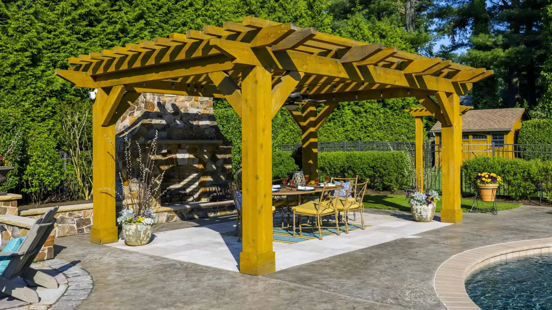 A wooden pergola graces the patio's seating area, next to an outdoor fireplace amidst lush greenery. With chic furniture and planters, it's a dreamy outdoor living space in Delaware County, PA, complemented by a pool with clear blue water. Perfect for hardscaping services inspiration.