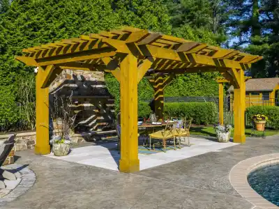 A wooden pergola graces the patio's seating area, next to an outdoor fireplace amidst lush greenery. With chic furniture and planters, it's a dreamy outdoor living space in Delaware County, PA, complemented by a pool with clear blue water. Perfect for hardscaping services inspiration.