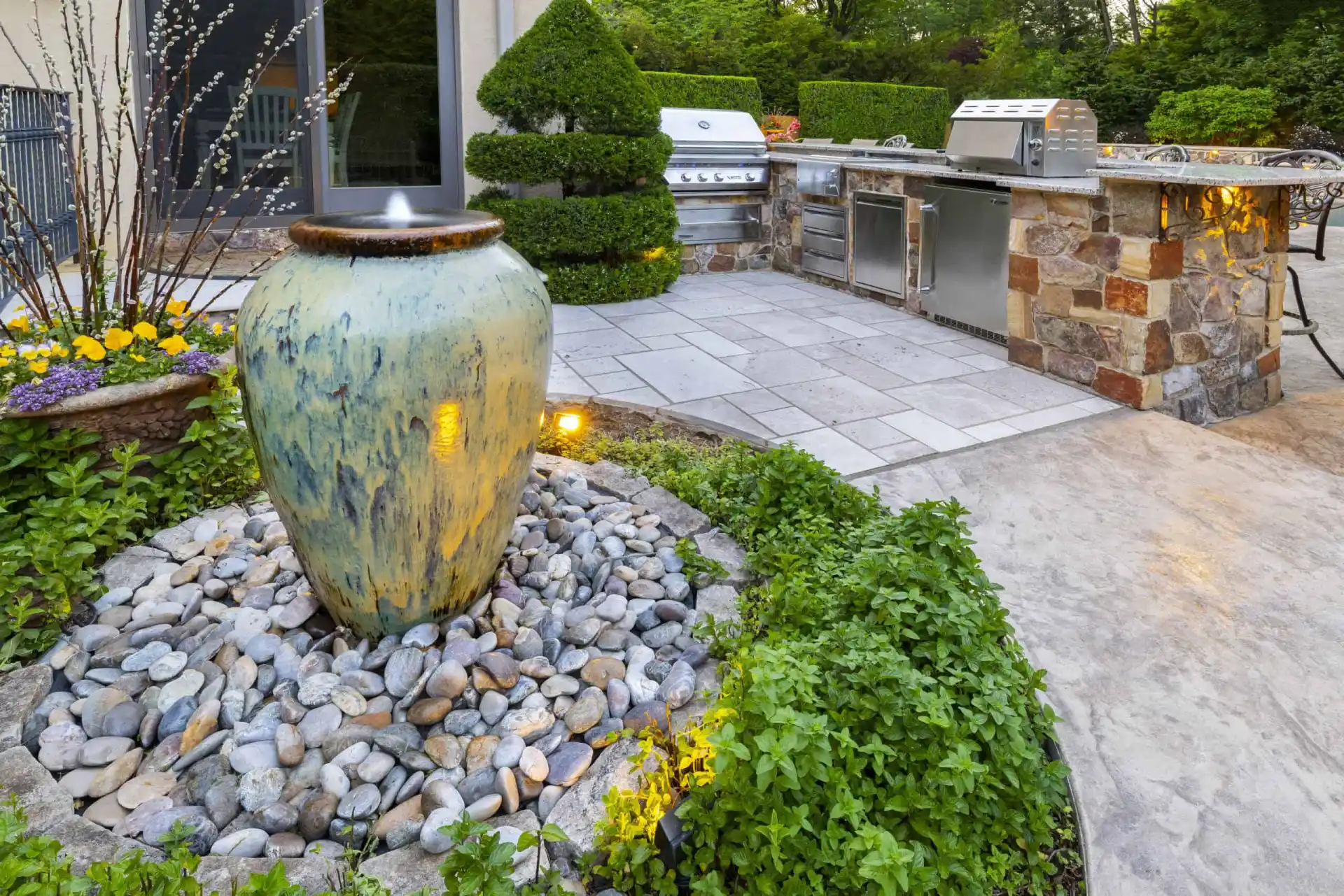 Nestled in Delaware County, PA, this outdoor living space features a patio adorned with a ceramic water feature, surrounded by lush plants and stones. An outdoor kitchen with stone countertops and stainless steel appliances stands ready for entertaining, enhanced by expert hardscaping services.