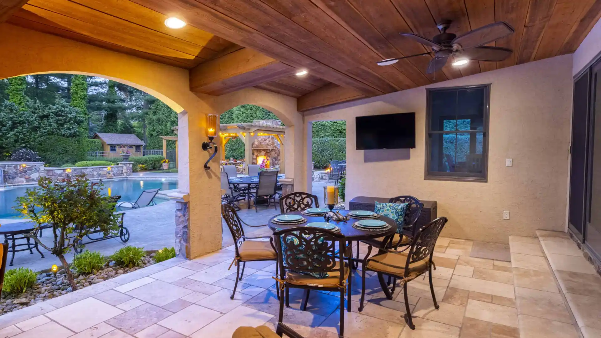 Covered patio with a black ceiling fan, wall-mounted TV, and round dining table for six. View through arches reveals a swimming pool and gazebo in a landscaped garden, crafted by expert landscaping services in Delaware County PA, all set against a backdrop of trees.