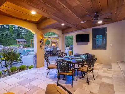 Covered patio with a black ceiling fan, wall-mounted TV, and round dining table for six. View through arches reveals a swimming pool and gazebo in a landscaped garden, crafted by expert landscaping services in Delaware County PA, all set against a backdrop of trees.