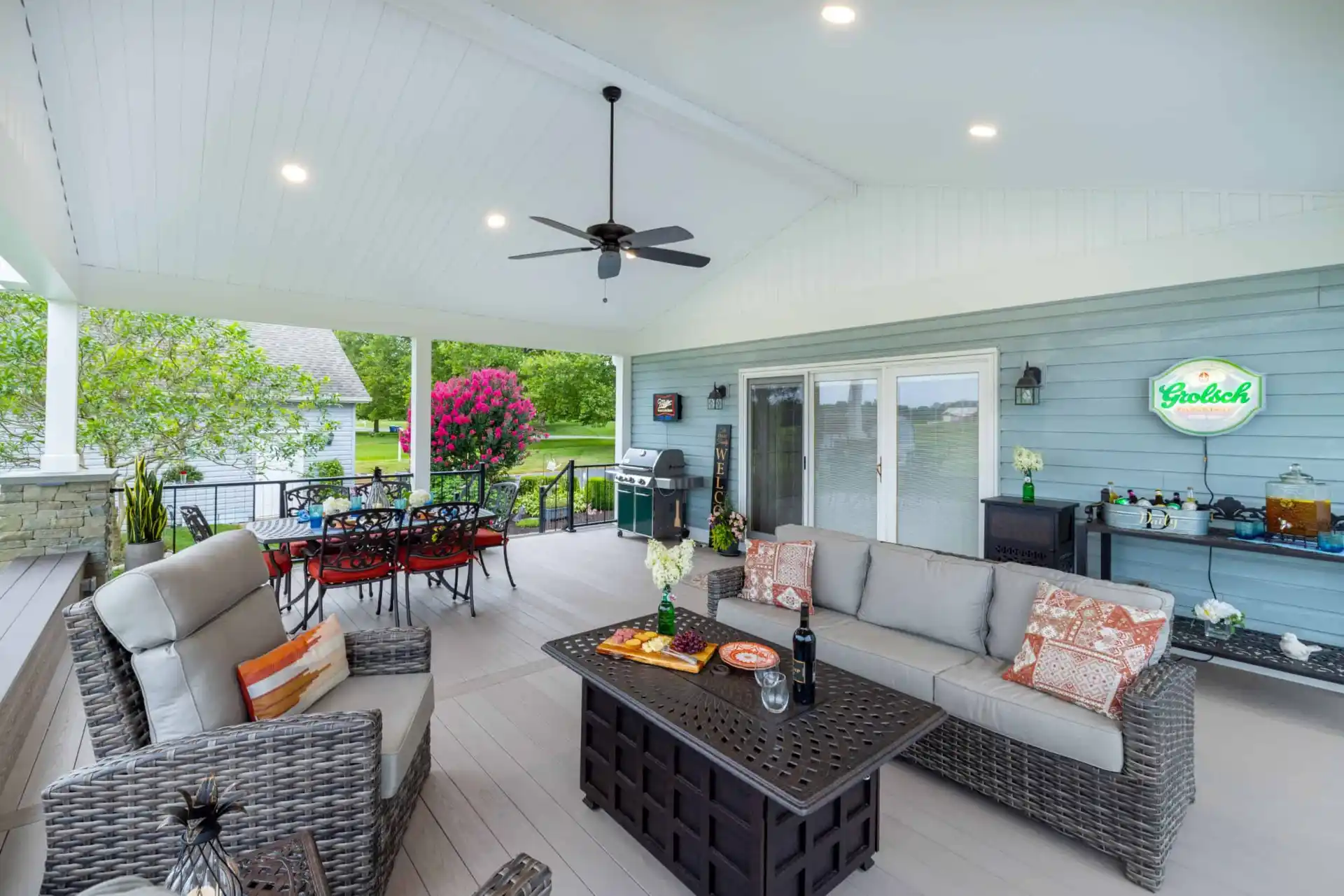 Spacious covered patio with wicker seating, a black metal table, ceiling fan, and outdoor kitchen. Vibrant pink flowers and greenery from top-notch landscaping services in Delaware County, PA surround the area. A neon 