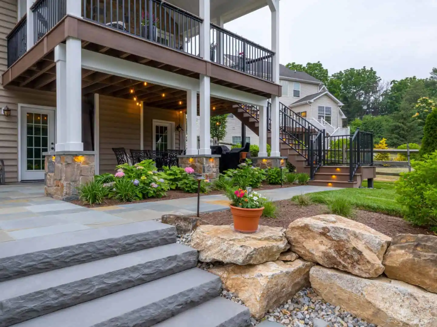 A two-story patio with a dining area beneath wooden decking showcases the best of outdoor living Delaware County PA. Steps, flanked by landscaped gardens with rocks and potted plants, lead up to the deck. Embedded lighting in the stairs illuminates this inviting space, with a house in the background.