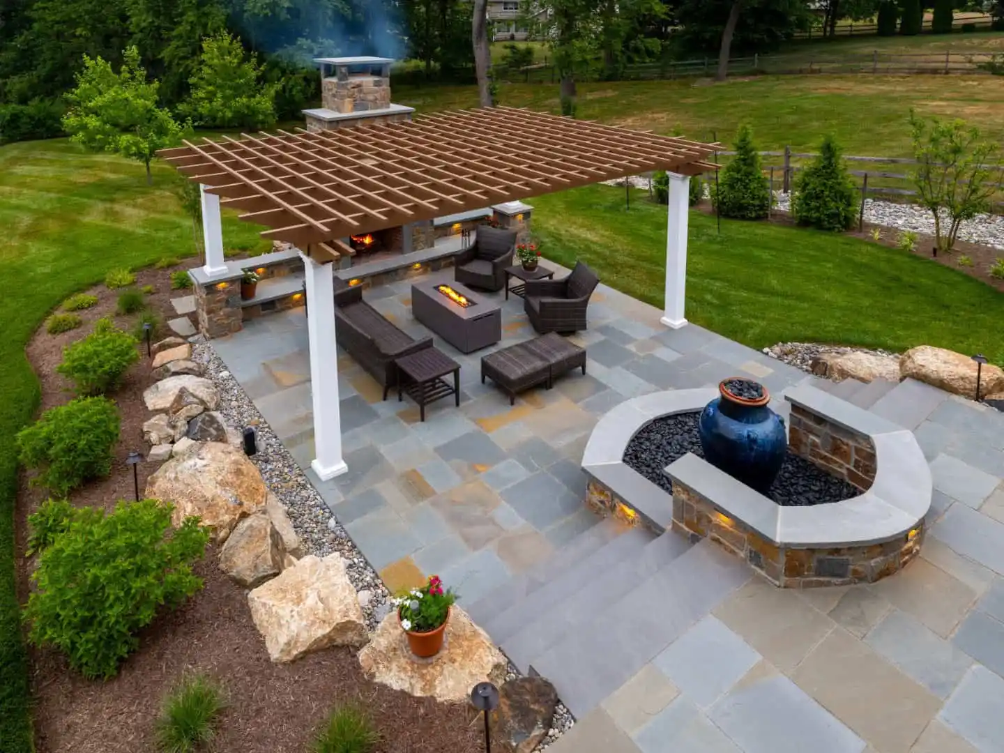 Aerial view of a landscaped backyard with a pergola-covered patio, featuring outdoor seating, a fireplace, and a fountain. Designed for optimal outdoor living in Delaware County, PA, the patio is surrounded by lush grass, trees, and flower beds with rocks.