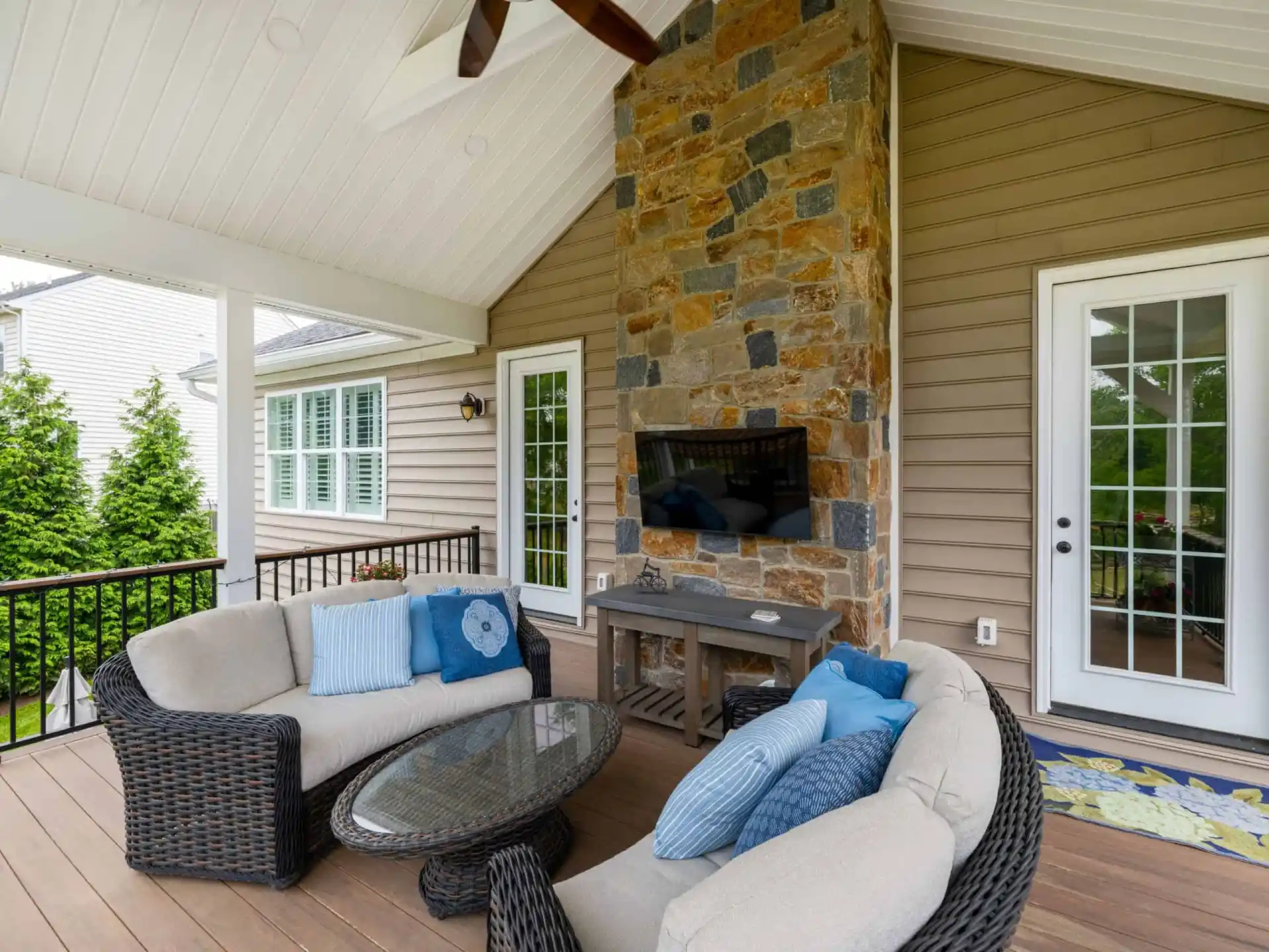 A covered patio with a vaulted ceiling in Delaware County, PA, featuring a stone fireplace, wall-mounted TV, and two wicker sofas with blue cushions. An oval glass coffee table sits at the center. White French doors lead inside while expert landscaping services bring the outdoor greenery to life.
