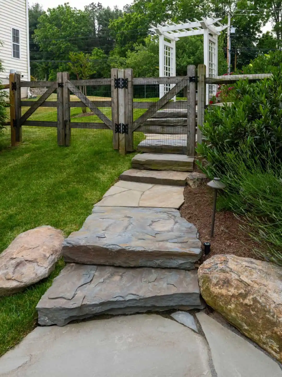 Stone steps lead up a grassy slope to a wooden garden gate, an inviting entryway designed with expert landscaping services in Delaware County, PA. A white pergola graces the background, while the path is lined with large rocks and lush green plants, enhancing your outdoor living experience.