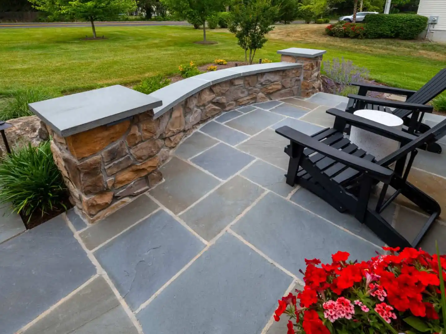 Stone patio with dark wooden chairs and a small round table. A stone wall with a curved design borders the patio, showcasing exceptional hardscaping services in Delaware County, PA. Red flowers in the foreground and a lush green lawn with trees add to the serene outdoor living experience.