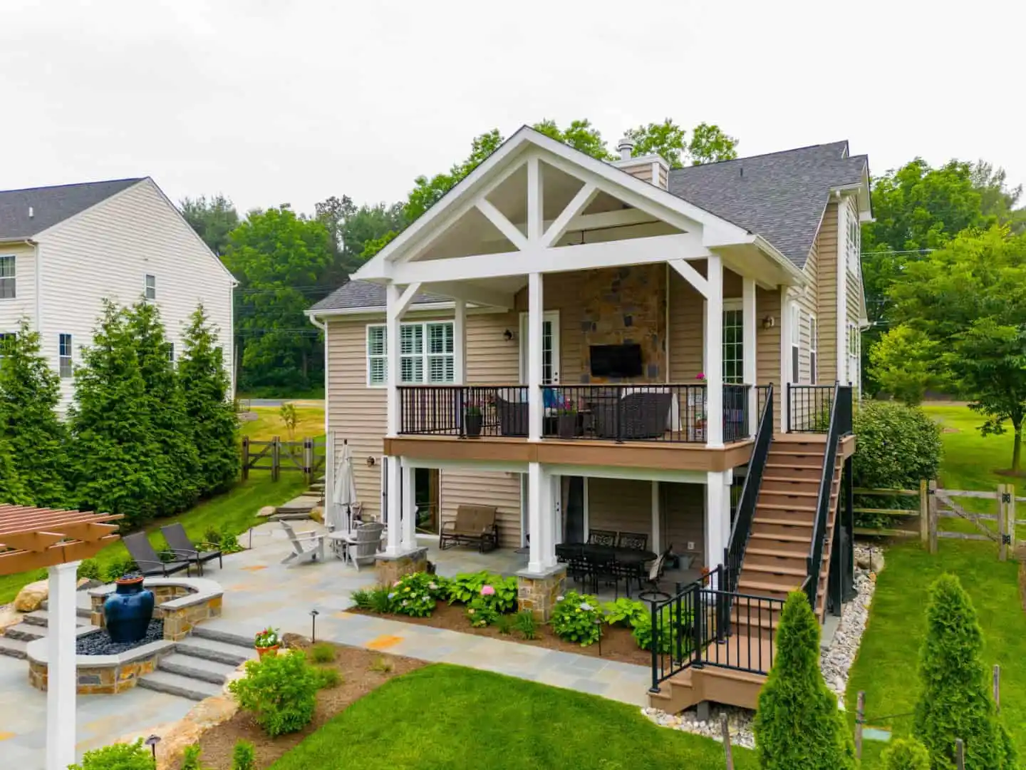 A two-story house in Delaware County, PA offers luxurious outdoor living. The spacious patio boasts a top deck with seating and TV, while below, lounge chairs circle a stone fire pit. Expert landscaping services adorn the yard with greenery and trees, complemented by a wooden staircase connecting the levels.