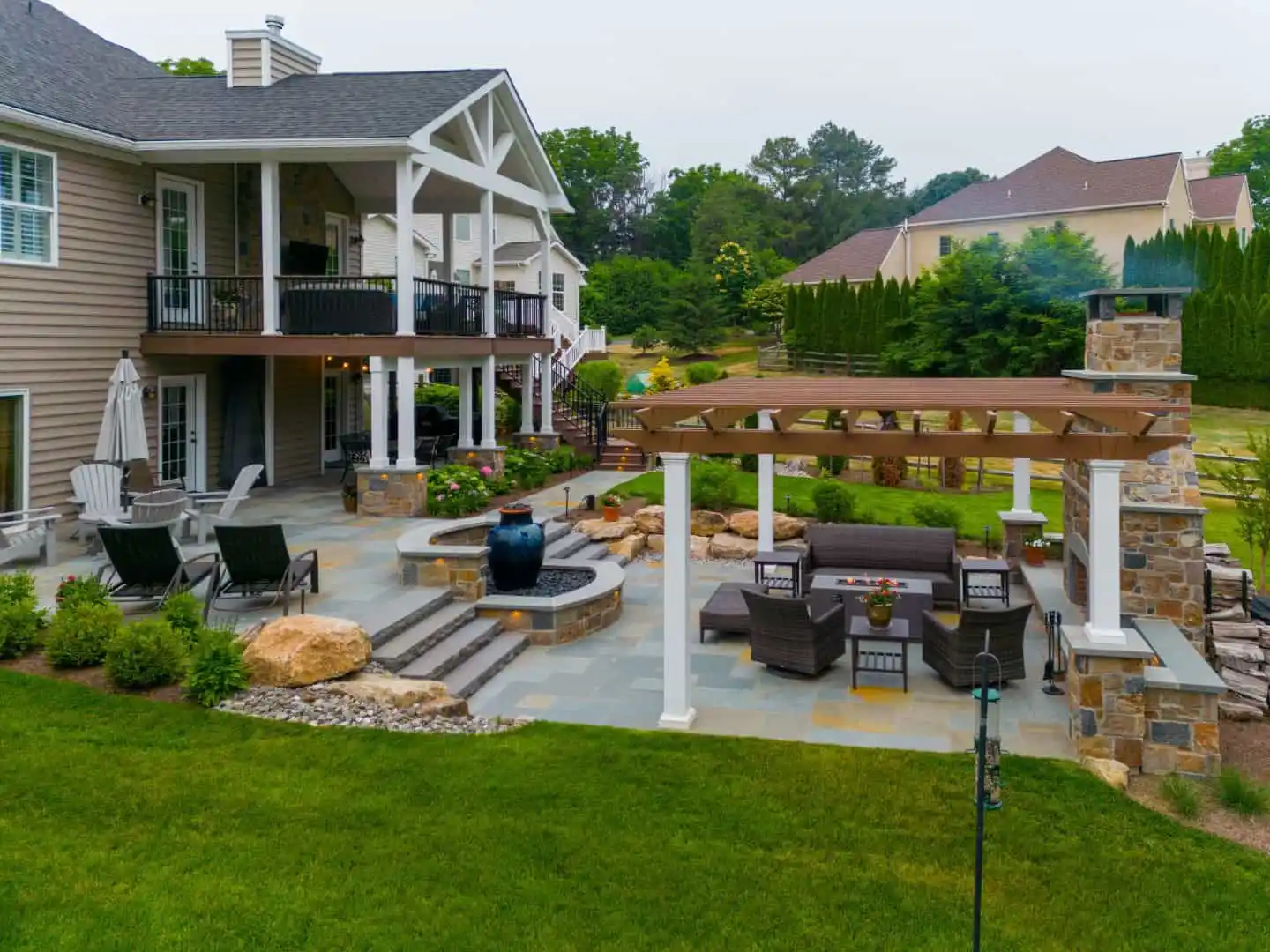 A modern backyard showcasing outdoor living in Delaware County, PA boasts a spacious patio with seating, a pergola, and a fire pit. The area, expertly landscaped with rocks and shrubs, features hardscaping services and includes a water fountain next to the two-story house with balcony furniture.