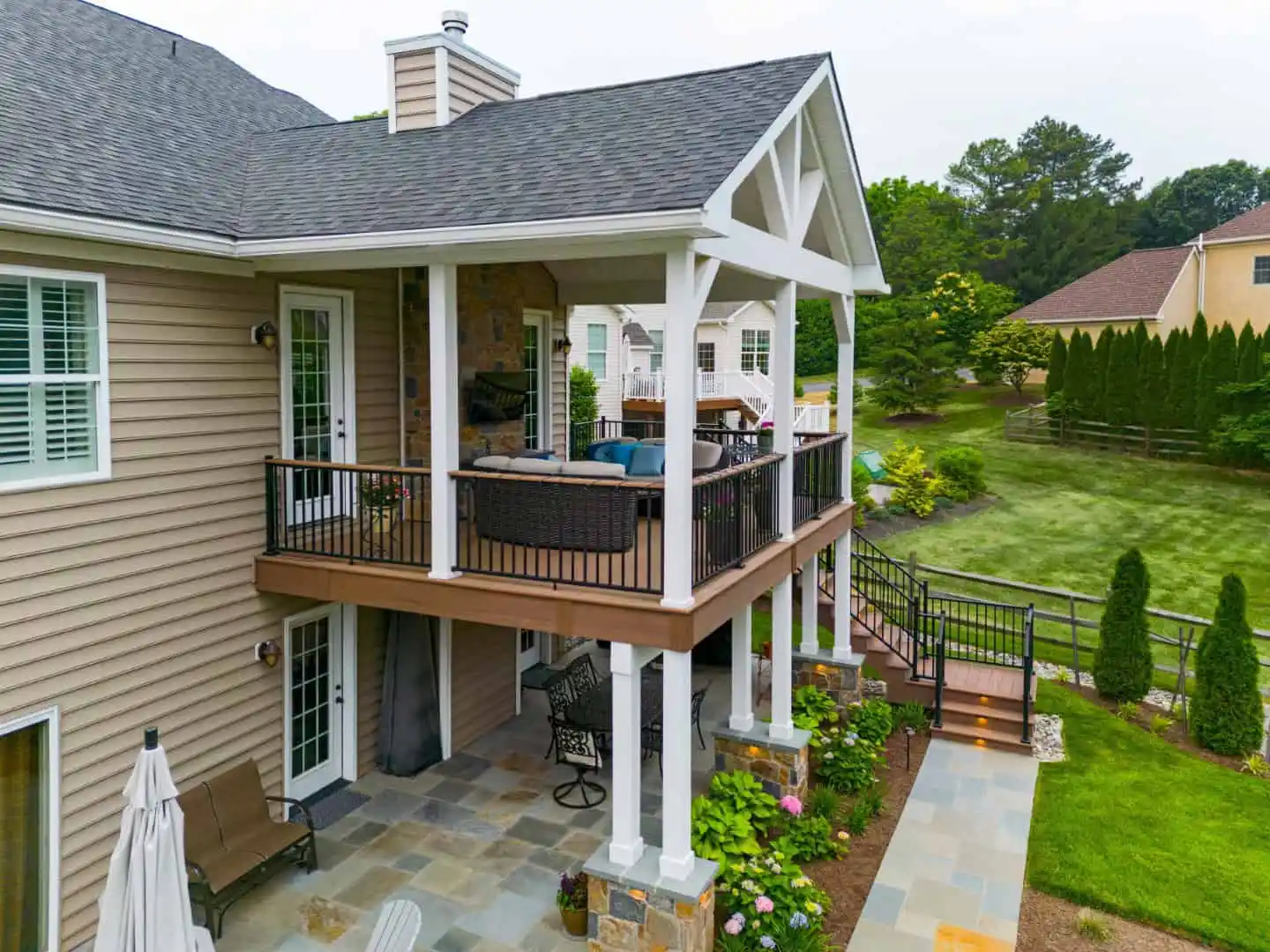 A two-story house with a grey roof and covered balcony showcases exquisite outdoor living in Delaware County, PA. The porch features inviting furniture, while the yard boasts lush green grass, elegant bushes, and trees. Stone and siding grace the exterior, complemented by a staircase leading to the balcony.