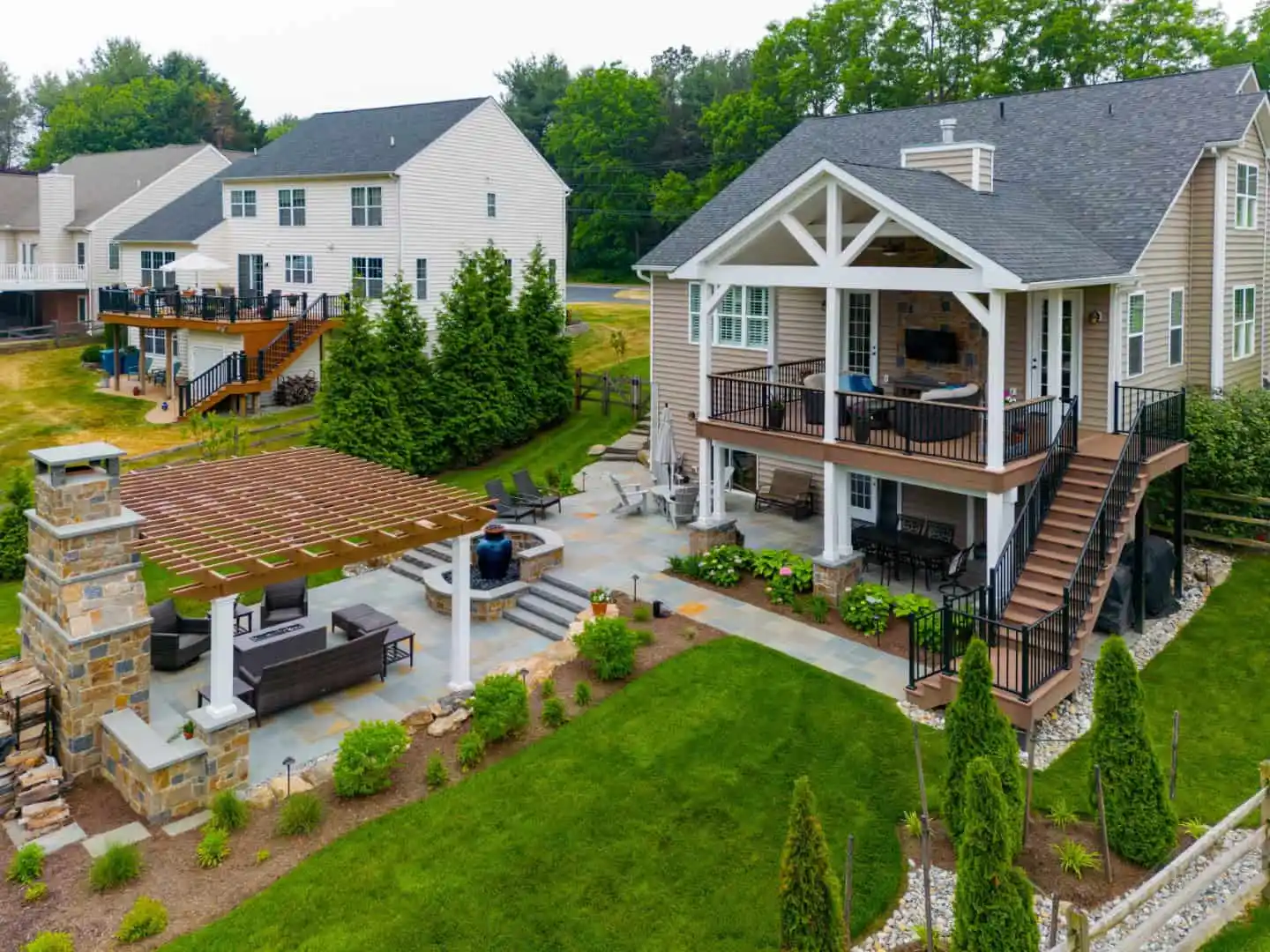 Aerial view of a modern suburban house in Delaware County, PA, with a spacious deck and covered patio. The backyard, crafted by expert landscaping services, features a stone fireplace, pergola, and well-maintained garden. Adjacent houses and lush green trees surround the area.