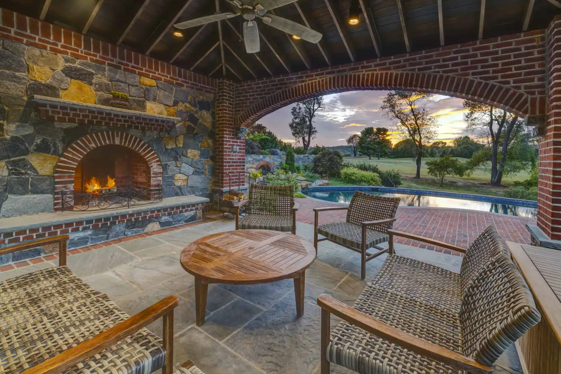 Covered patio with wicker chairs and a round wooden table, stone fireplace with a fire burning, brick arches framing a view of a pool and trees at sunset in the background.