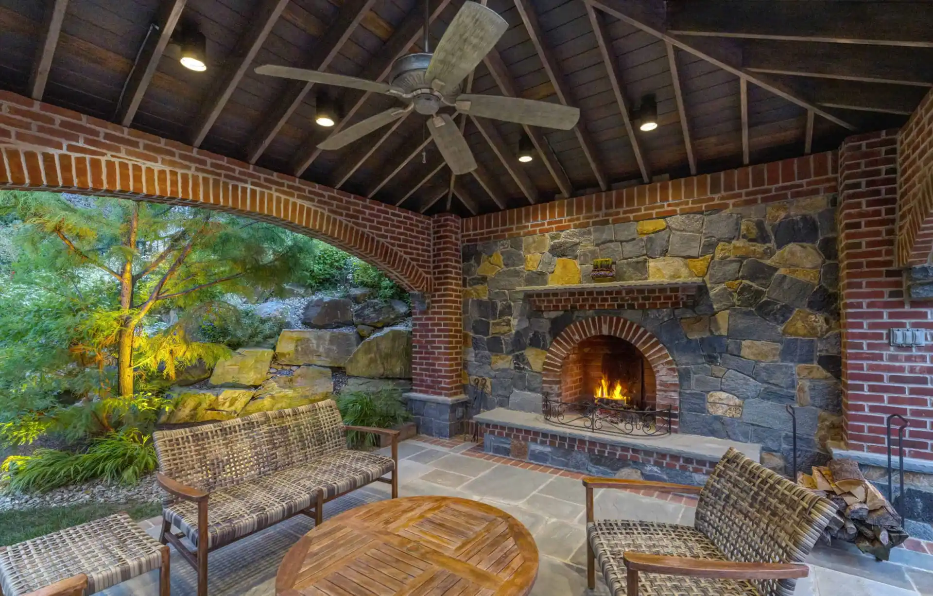 Covered patio with brick and stone fireplace, wooden ceiling with fan, wicker seating, round wooden table, and view of lush garden through large arched opening.
