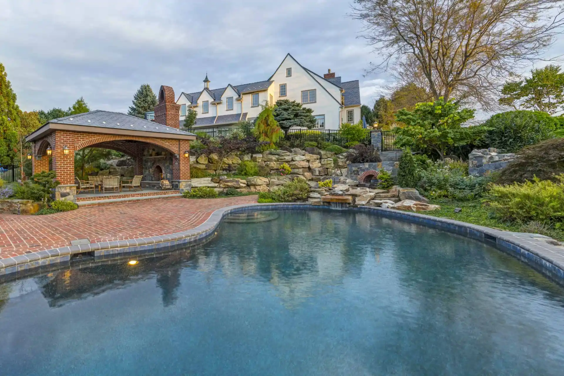 A large house with white walls and dark roof sits atop a landscaped hill with lush greenery, rock features, and a brick-covered gazebo next to a curved swimming pool under a cloudy sky.