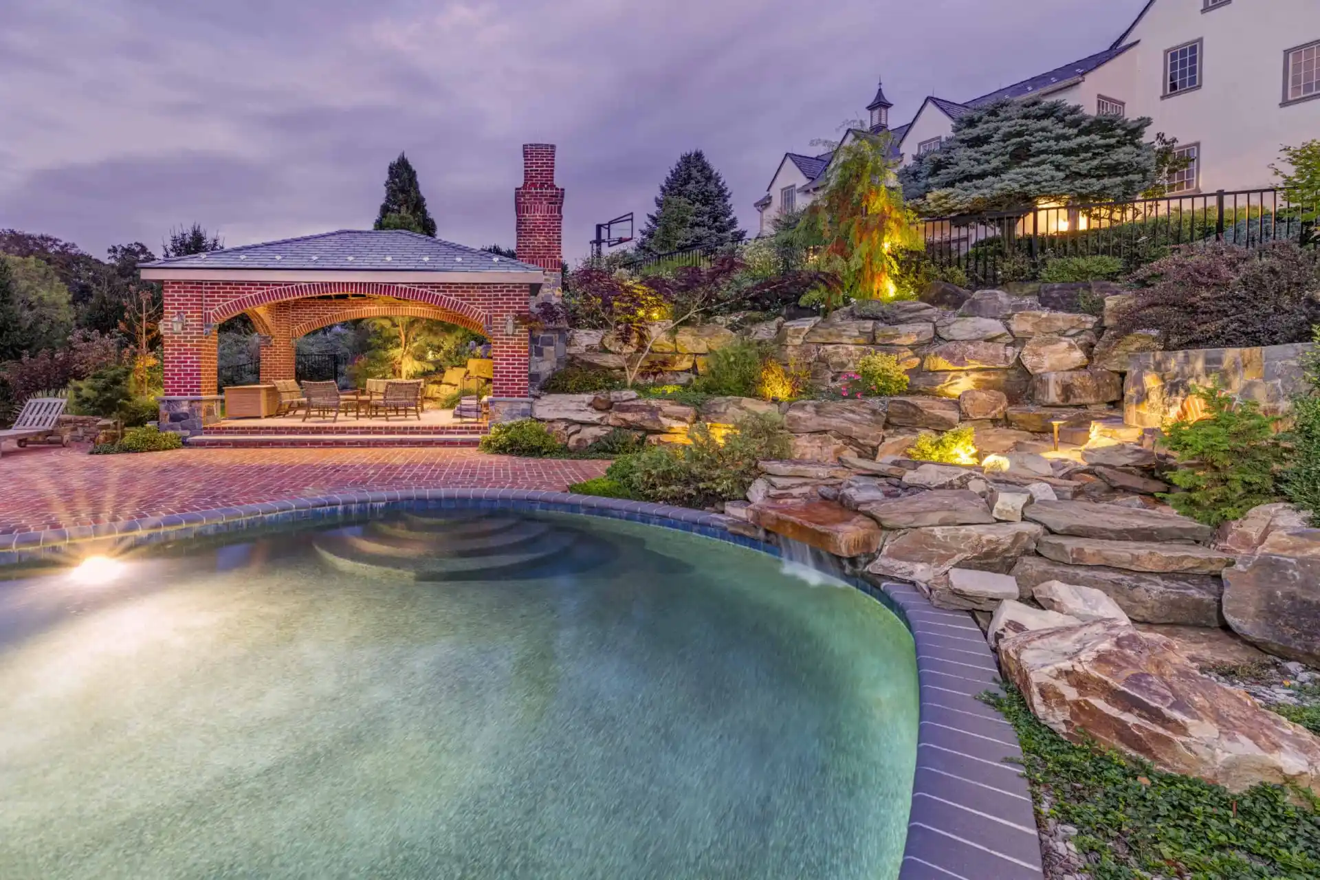 Luxurious backyard with a curved pool, stone waterfall feature, and illuminated landscaping. A brick patio with a covered seating area and fireplace is surrounded by lush trees and shrubbery near a large house at dusk.