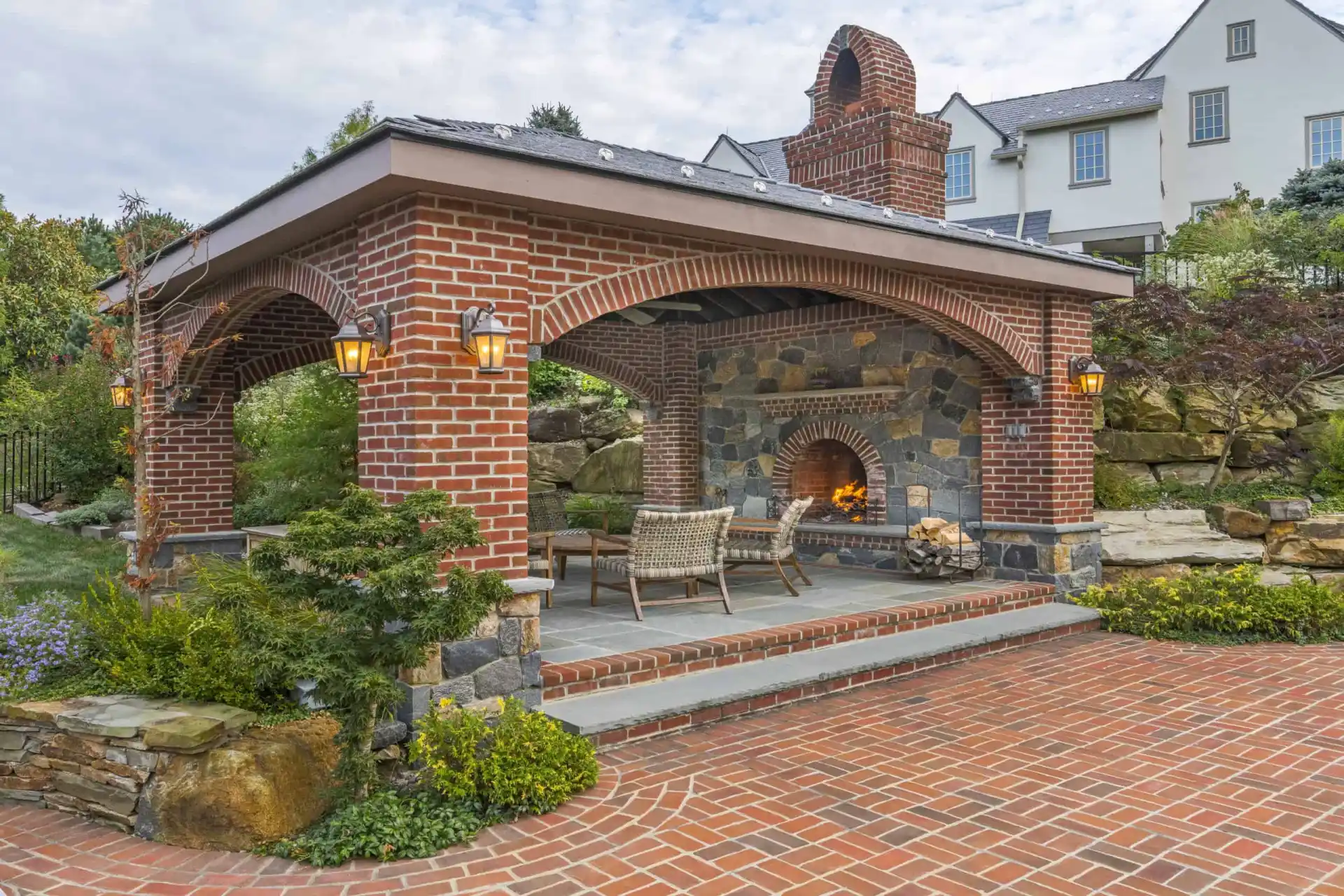 A covered outdoor patio with brick arches, stone fireplace, and two chairs, surrounded by greenery and brick paving, with a large house and landscaped garden in the background.