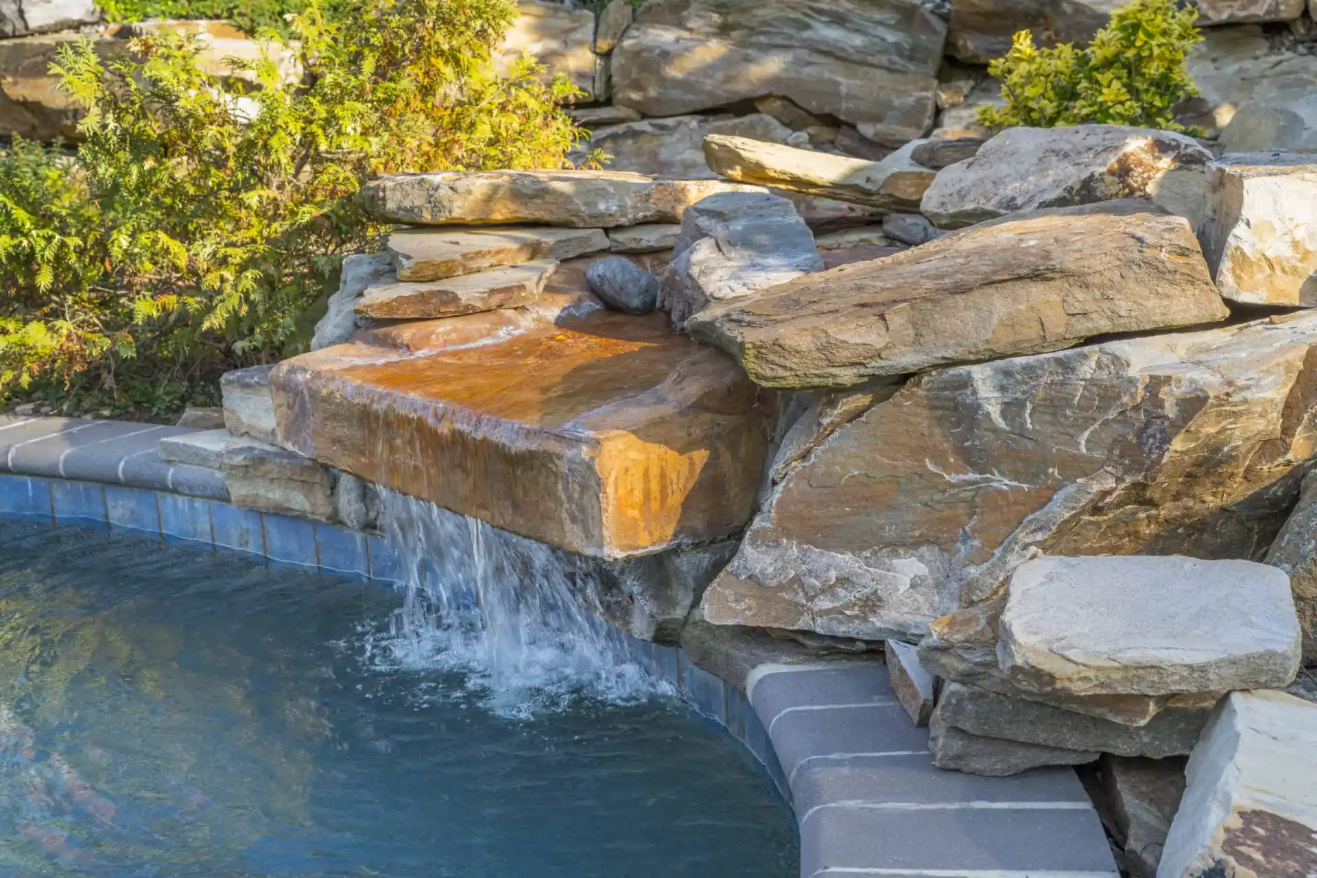 Water cascades from a flat stone feature built into a pile of large, natural rocks into a swimming pool, surrounded by greenery and tile edging.