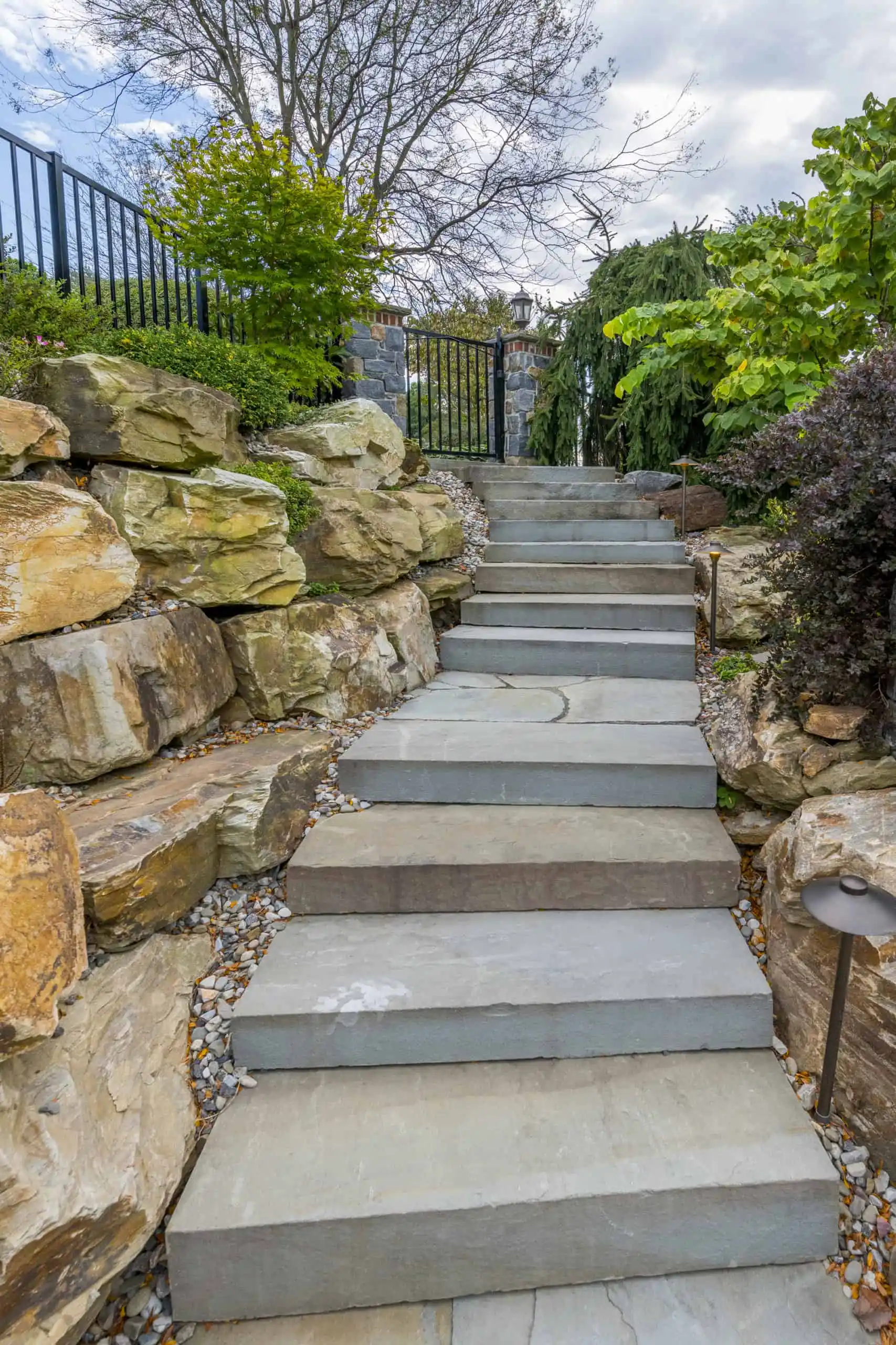 Stone steps lead up through a landscaped garden with large rocks, green bushes, and a black metal fence at the top, under a cloudy sky.