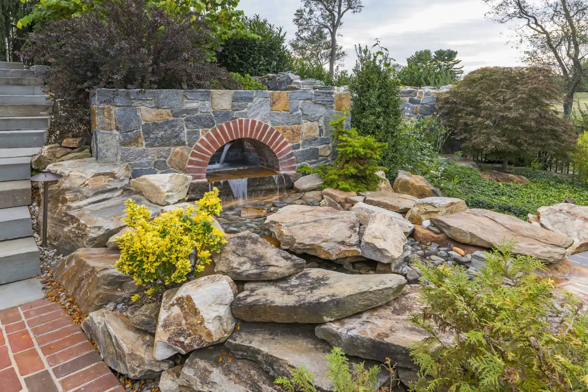 A small waterfall flows from a stone wall with a brick arch into a rock garden, surrounded by various green shrubs and plants, beside a stone staircase and brick pathway.