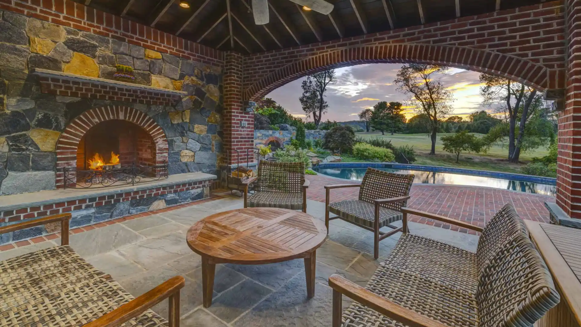 Experience the ultimate in outdoor living in Delaware County, PA, with a stone fireplace illuminating wicker furniture. This charming patio overlooks a pool and open field with sunset views through an arched brick opening, enhanced by a ceiling fan and wooden beams.