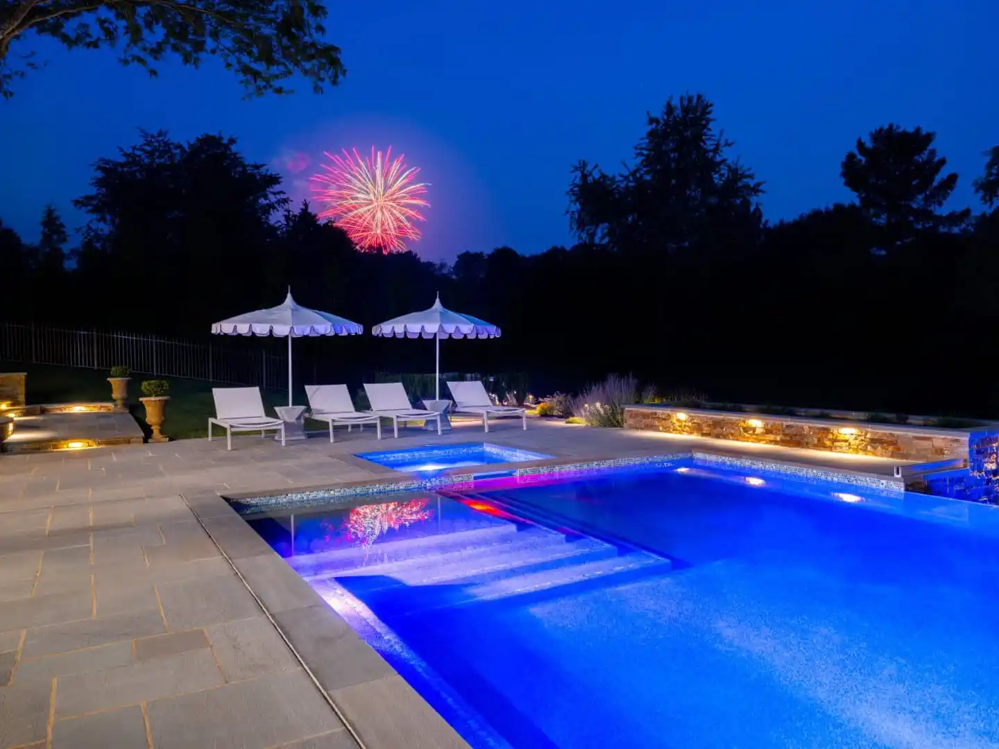A large, illuminated swimming pool at night with lounge chairs and umbrellas to the side showcases stunning outdoor living. Fireworks burst in the sky behind silhouetted trees in Delaware County, PA, adding vibrant colors to a meticulously designed landscape.