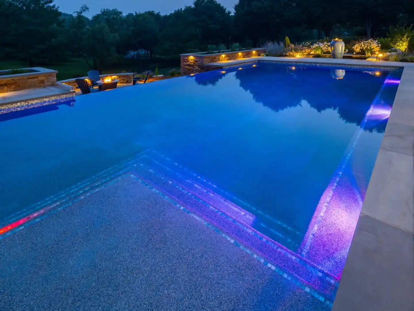 A tranquil swimming pool at night features glowing blue and purple underwater lights. Surrounded by elegant landscape lighting and trees under a starry sky, it offers a serene escape. It's an inviting outdoor living space in Delaware County, PA's finest landscaping tradition.