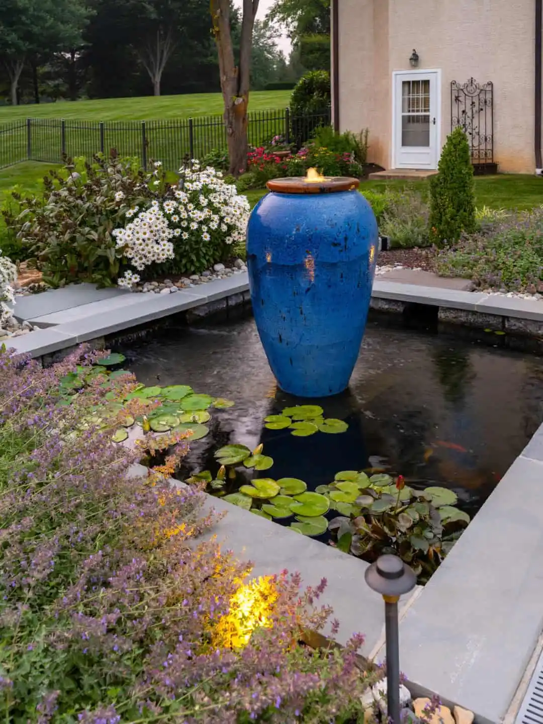 A garden pond with lily pads features a tall, blue ceramic fountain vase at its center. The surrounding landscaping services in Delaware County, PA include flowering bushes, small trees, and a neatly trimmed lawn. A stone path leads to a nearby building with a white door.