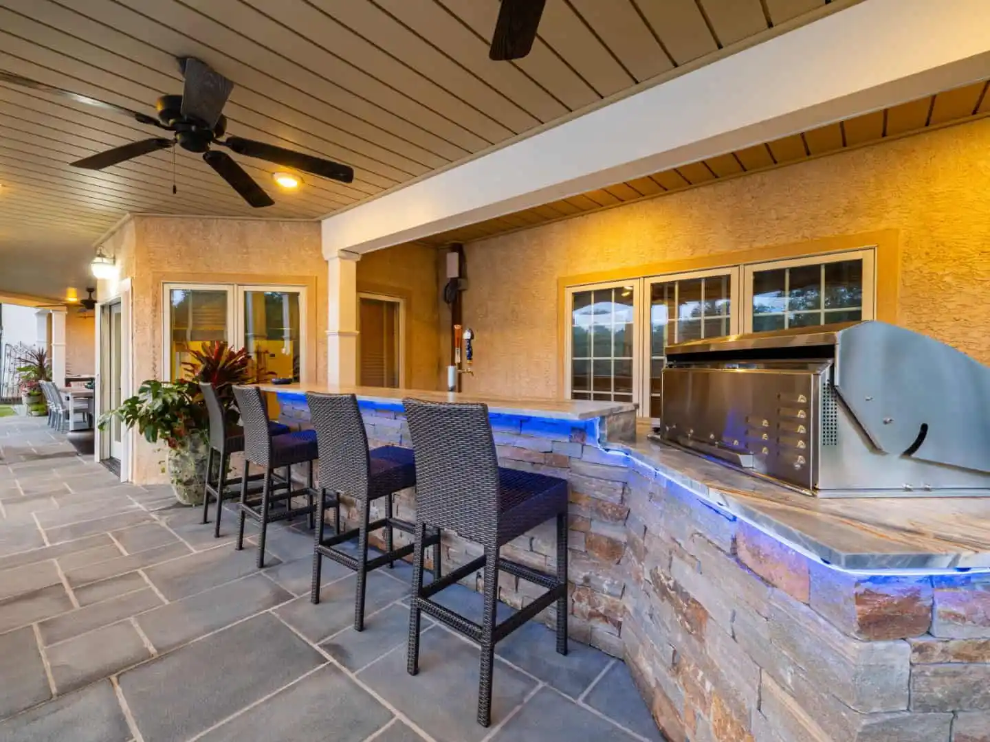 Outdoor living in Delaware County, PA is elevated with a stone bar counter and three woven bar stools. A grill rests under the wooden ceiling, complete with fans. The warm glow of recessed lighting and lush potted plants complete this inviting oasis.