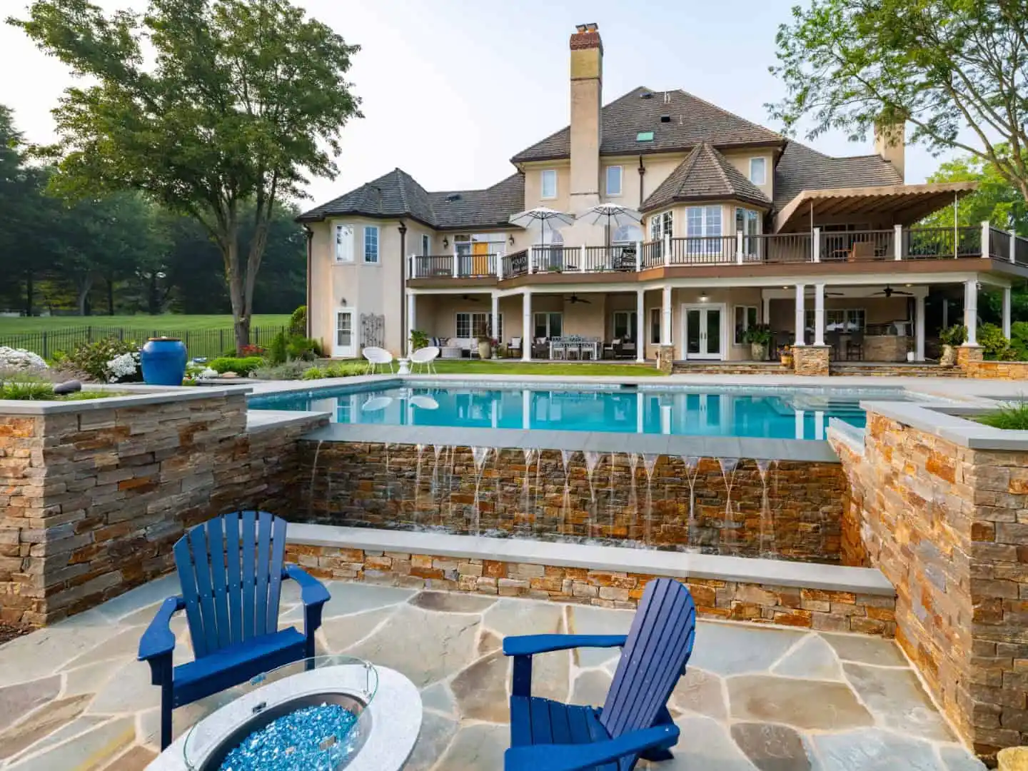 Luxurious house with stone patio features a multi-level pool and waterfall. Blue Adirondack chairs surround a fire pit. The house, enhanced by expert hardscaping services in Delaware County, PA, boasts large windows, balconies, and a spacious outdoor dining area amid lush greenery.