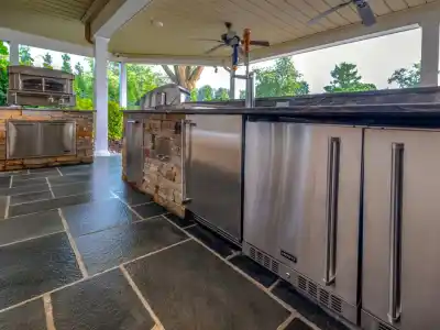 Outdoor living in Delaware County, PA, is epitomized by this kitchen, boasting stainless steel appliances and stone accents. A marble countertop complements an oven, fridge, and tap on a slate tile floor with lush greenery beyond. Ideal for those seeking exquisite hardscaping services.