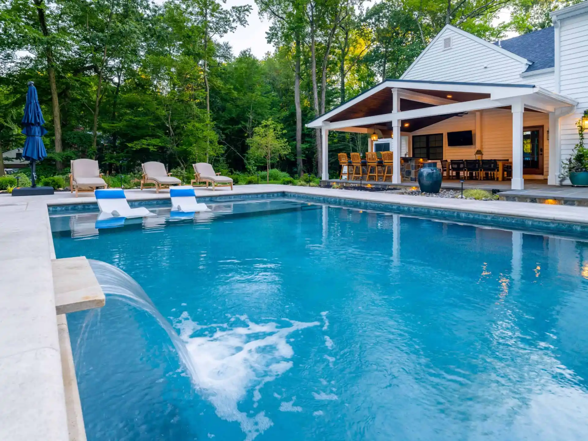 A luxurious backyard with a large pool featuring water fountains is a dream of outdoor living in Delaware County, PA. Three lounge chairs sit poolside, and in the background, a modern white house with a covered patio area and bar stools is surrounded by lush green trees.